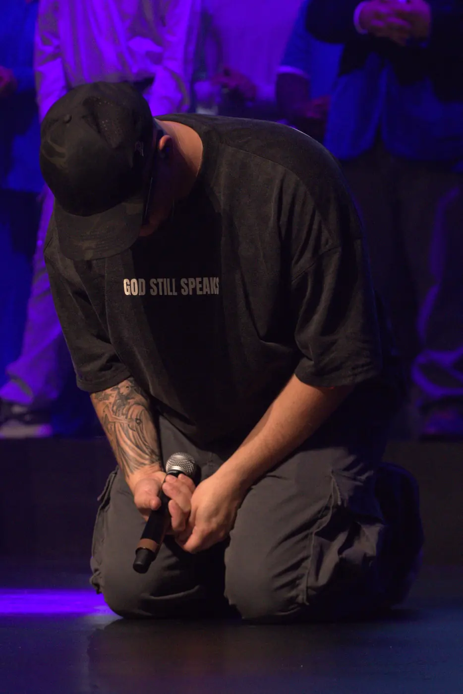 A man kneeling in prayer in a GOD STILL SPEAKS shirt